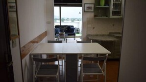 Uma área de refeições com uma mesa branca e quatro cadeiras. Vê-se um varanda pela janela.