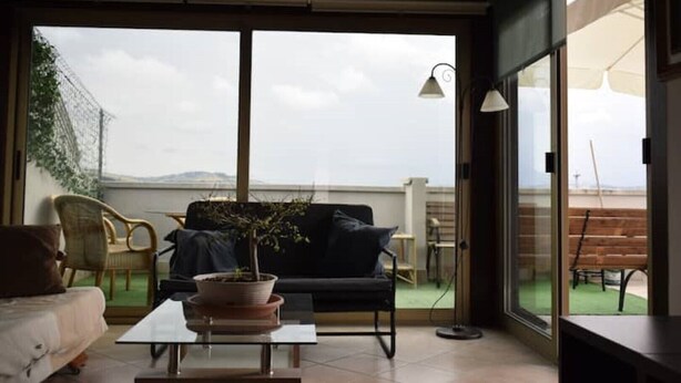 Quarto com sofá, mesa de centro e planta em vaso. Há uma porta de vidro deslizante que dá para área externa com vista do céu e paisagem distante.