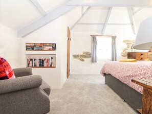 Ett sovrum med ett sluttande tak, en säng med ett blommigt överkast, en bokhylla fylld med böcker, en vägg med stenbeklädnad och ett fönster med gardiner.