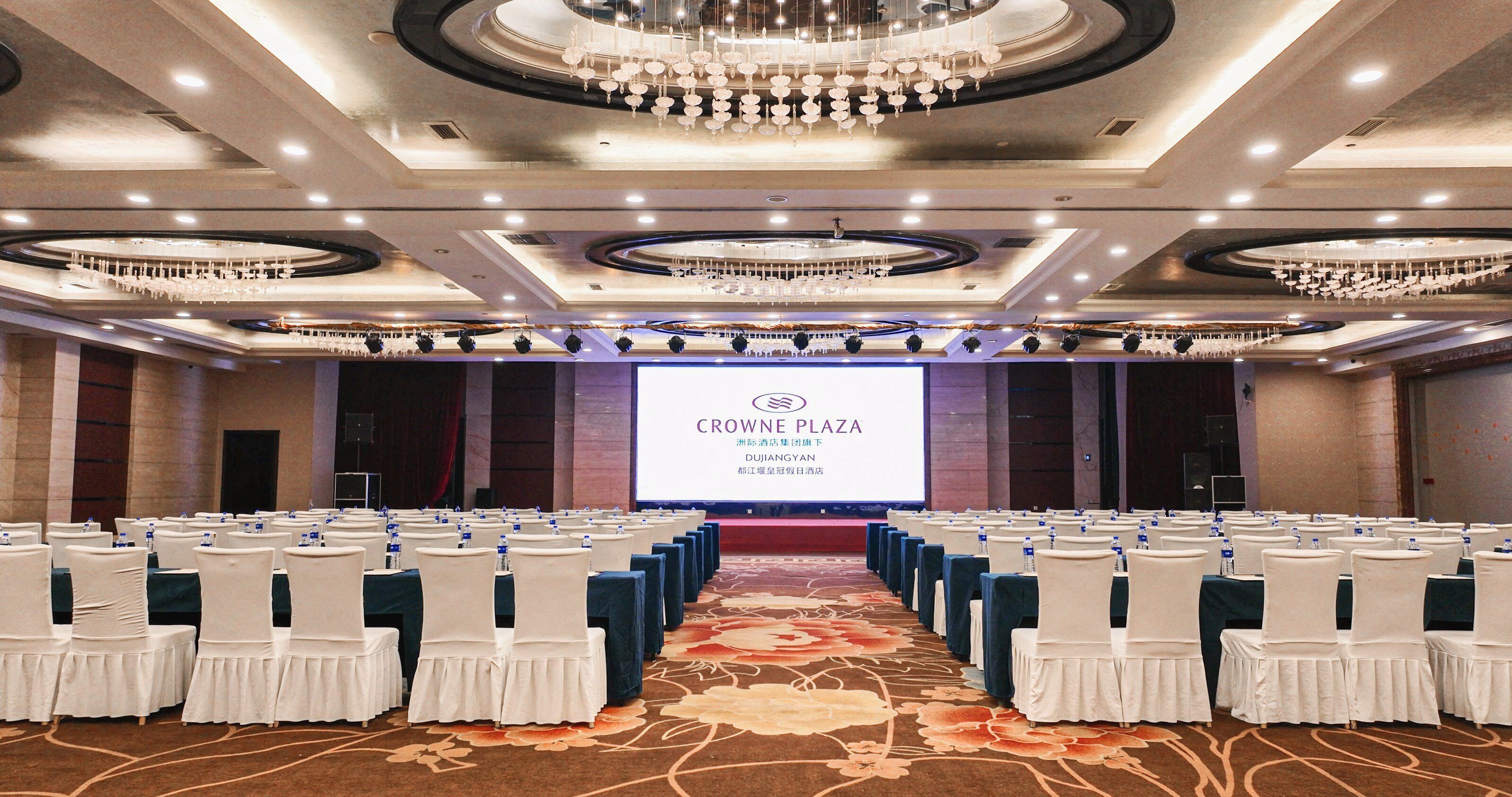 A conference room with rows of white chairs, a large screen displaying 'CROWNE PLAZA', and a chandelier.