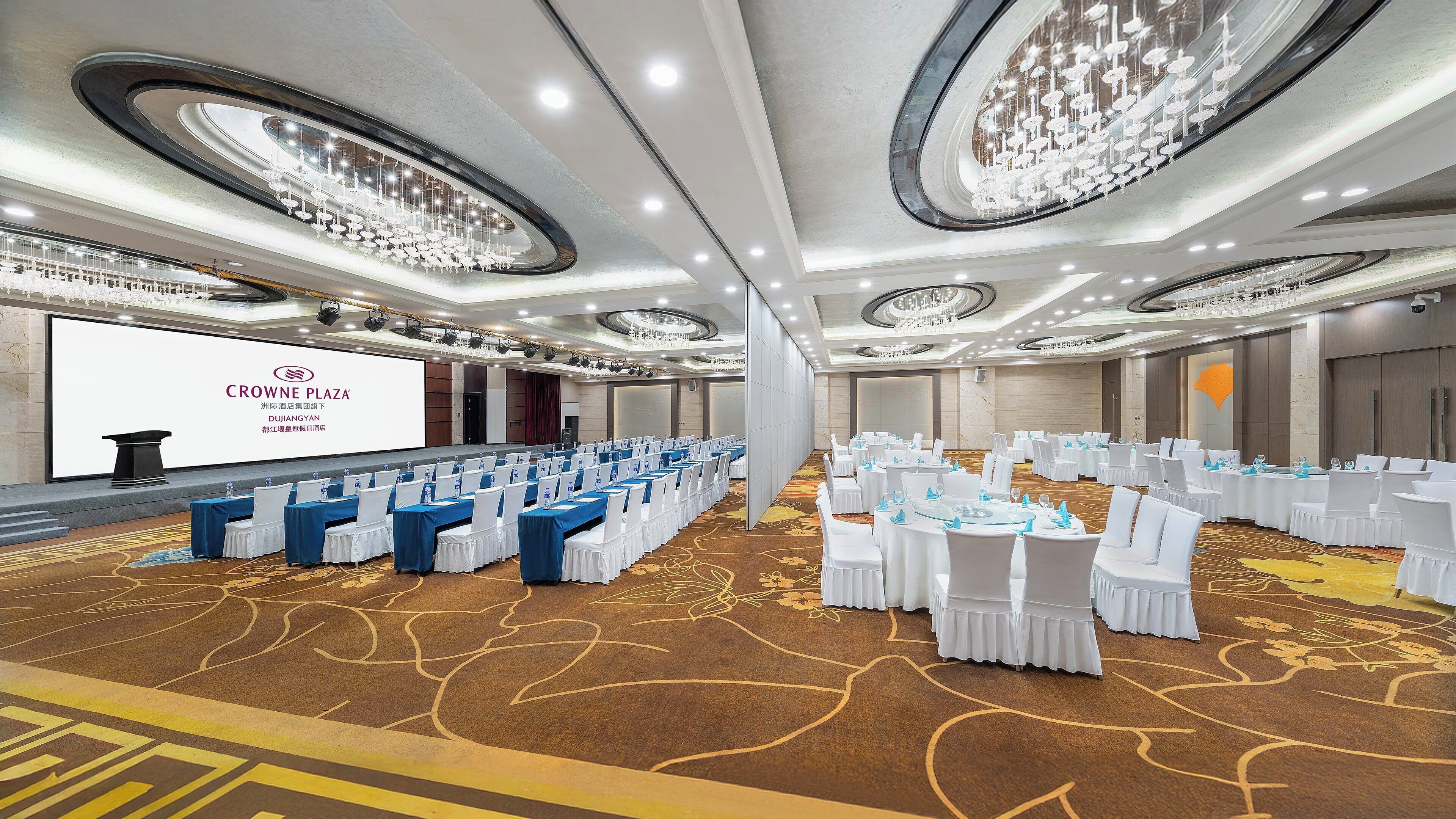A conference room with rows of tables, white chairs, and a large screen displaying text.