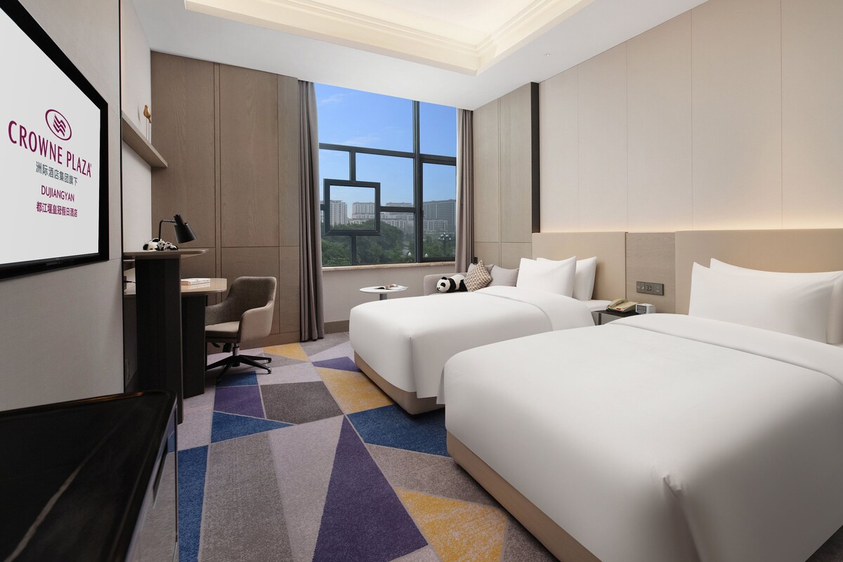 A hotel room with two beds, a desk, a chair, and a large window offering a city view.
