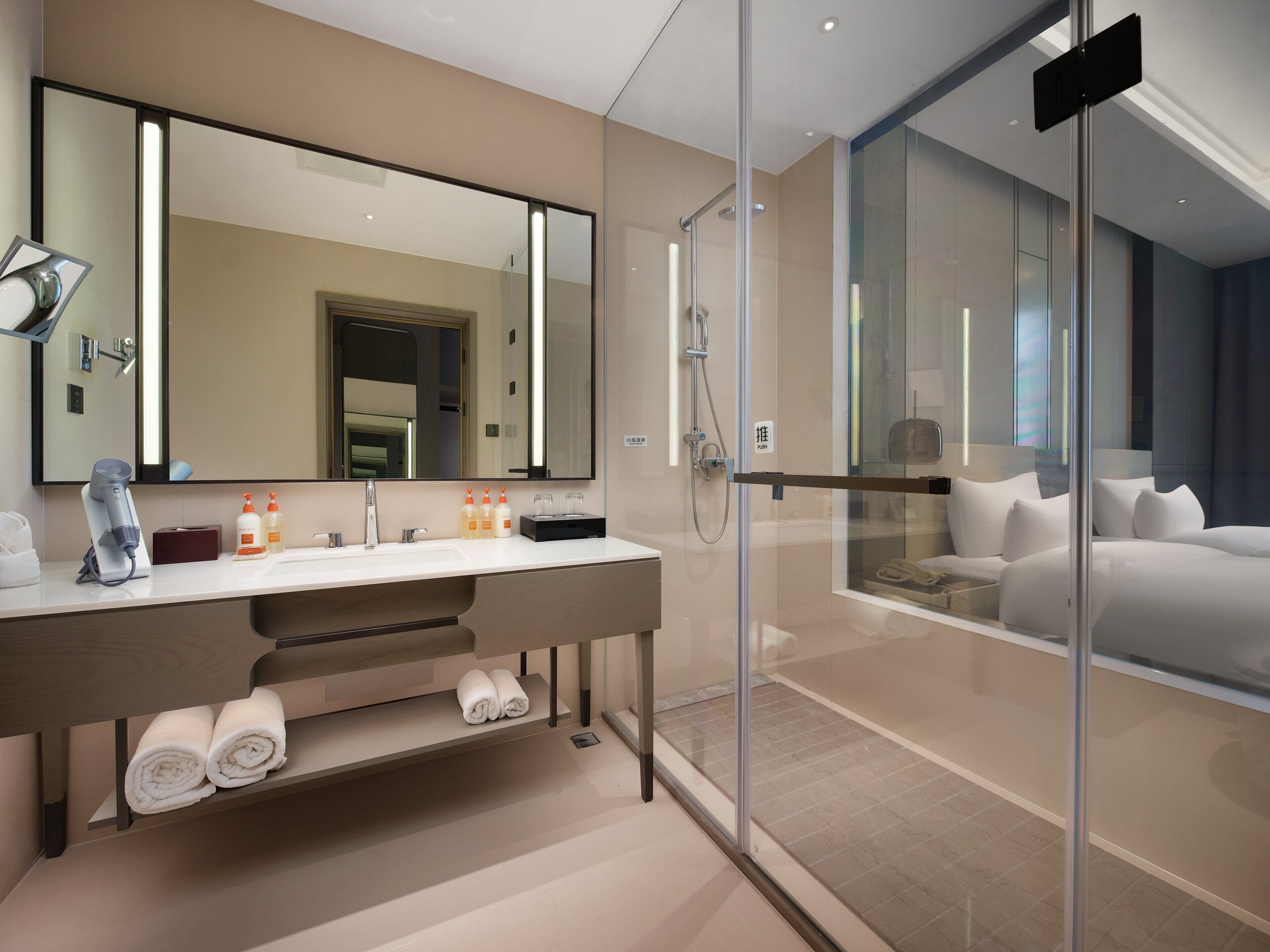 A modern hotel bathroom with a large mirror, a shower enclosure, and a bed visible through a glass partition.