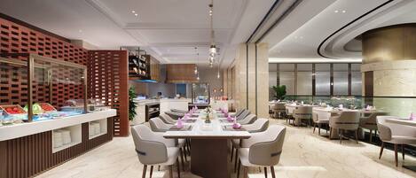 A modern dining area with a long table set for a meal, surrounded by chairs, and a kitchen area in the background.