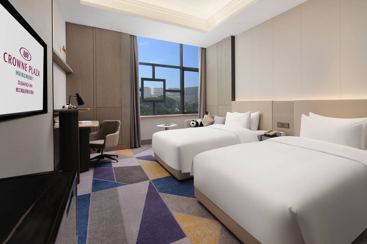 Habitación de hotel con dos camas, un escritorio, una silla y un amplio ventanal con vista a la ciudad.