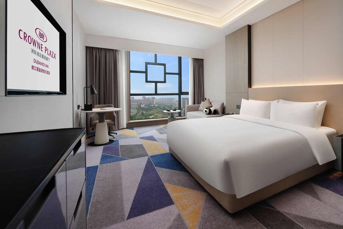 A modern hotel room with a large bed, a desk, a chair, and a view of the city.