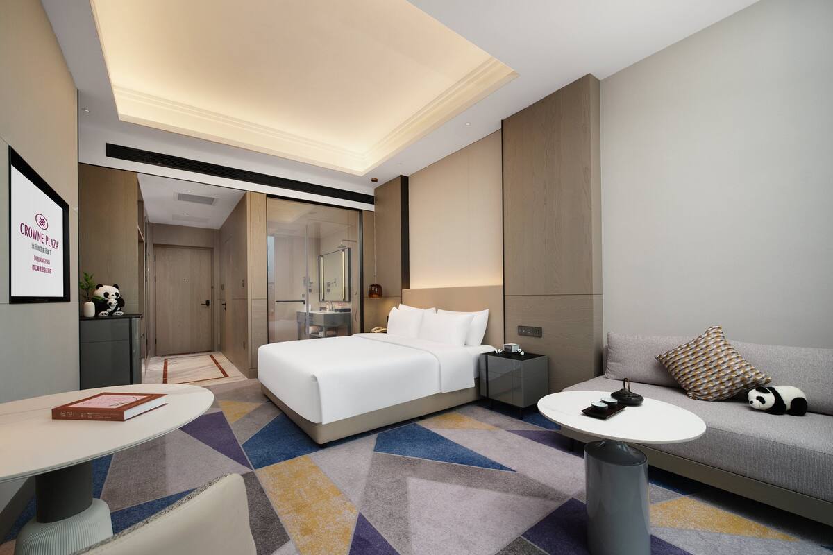 Una habitación de hotel moderna con una cama grande, un sofá, una mesita pequeña y una mesita de noche.