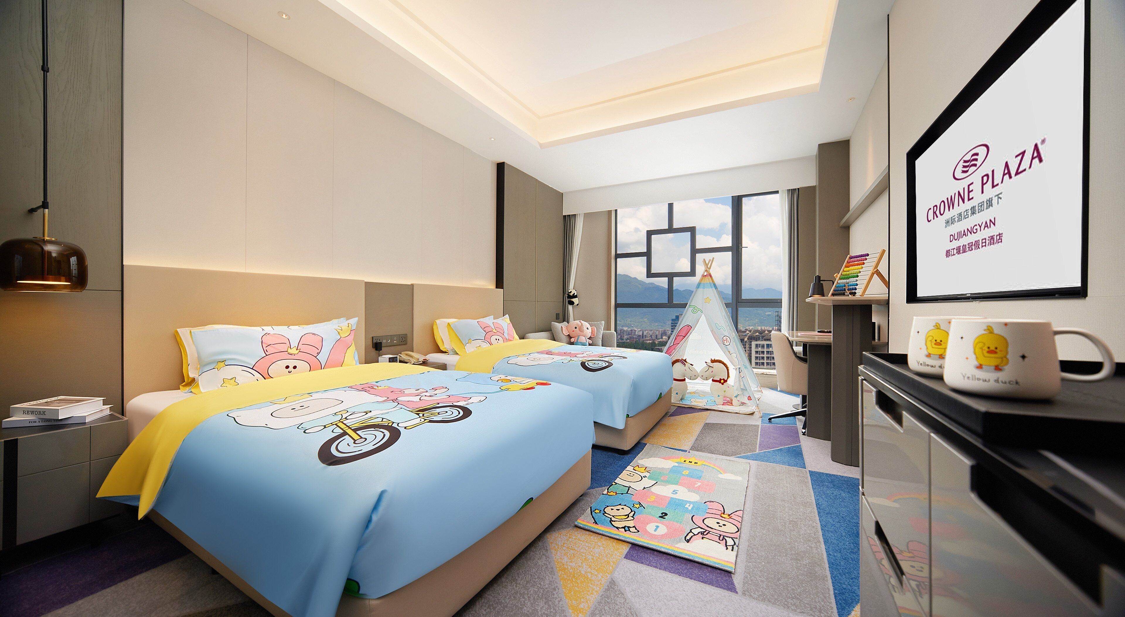 A hotel room with two beds, a desk, a chair, a window with a city view, and a sign for Crowne Plaza Dujang.