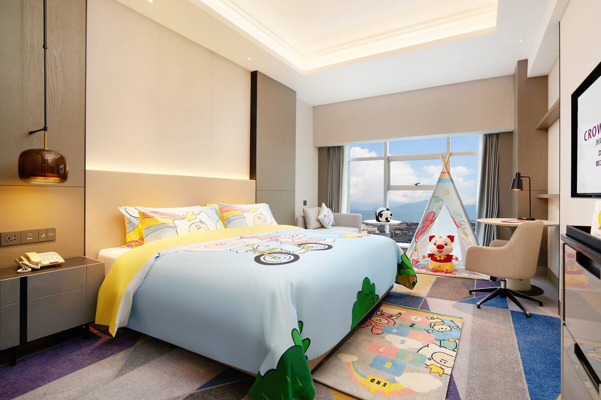 Una habitación de hotel con cama, escritorio, silla, ventana y pantalla que muestra el nombre del hotel.