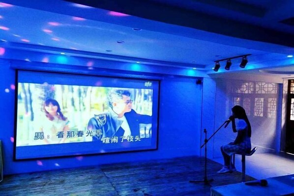 A room with a large screen displaying a movie scene, a microphone stand, and a person sitting on a chair.