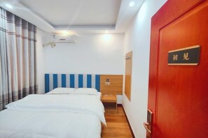 Ένα δωμάτιο ξενοδοχείου με ένα κρεβάτι, ένα ξύλινο κομοδίνο, μια κόκκινη πόρτα με μια επιγραφή και ένα παράθυρο με κουρτίνες.