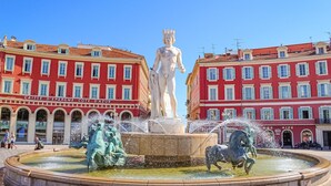 A large statue in a fountain with water jets, surrounded by a circular pool, set against red buildings with balconies and windows. - One minute from Place Masséna,  fully equipped STUDIO with balcony (Nice)