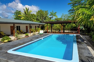 Una piscina rectangular con borde de azulejos, rodeada por un patio con sillas y plantas en macetas.