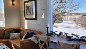 A cozy living room with a wooden floor, a brown leather sofa, a dining table with two wooden chairs, and a large window with a view of a snowy landscape.