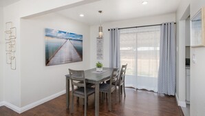 A dining area with a wooden table and chairs, a painting on the wall, and a window with curtains.