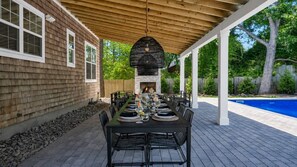 Patio esterno con un lungo tavolo da pranzo apparecchiato per un pasto, un camino in pietra e una piscina sullo sfondo.
