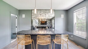 Une cuisine moderne dotée d'un grand îlot central, de trois chaises en osier, de luminaires suspendus et offrant une vue sur l'extérieur grâce à une fenêtre.