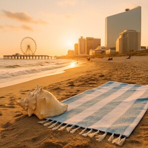 A beach scene with a striped beach towel and a seashell on the sand, a pier with a Ferris wheel in the background, and high-rise buildings. - 3BR Near AC Hospital – All Utilities Included – Travel Nurse Friendly (Atlantic City)