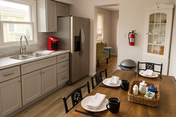 A modern kitchen with stainless steel appliances, a dining area with a wooden table set for two, and a small dining table with a basket of condiments. - 3BR Near AC Hospital – All Utilities Included – Travel Nurse Friendly (Atlantic City)