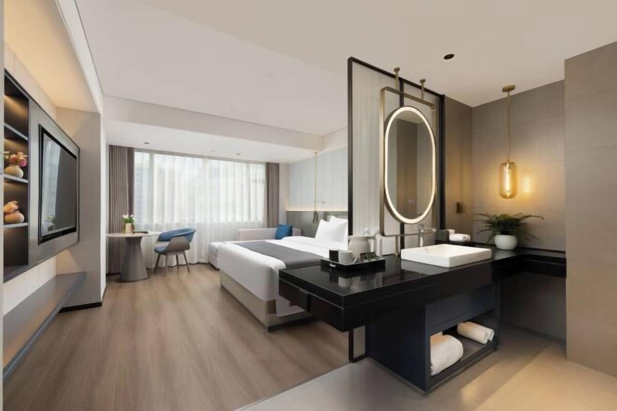 A modern hotel room with a large bed, a desk with a mirror, a separate bathroom with a vanity, and a seating area with a chair and table.
