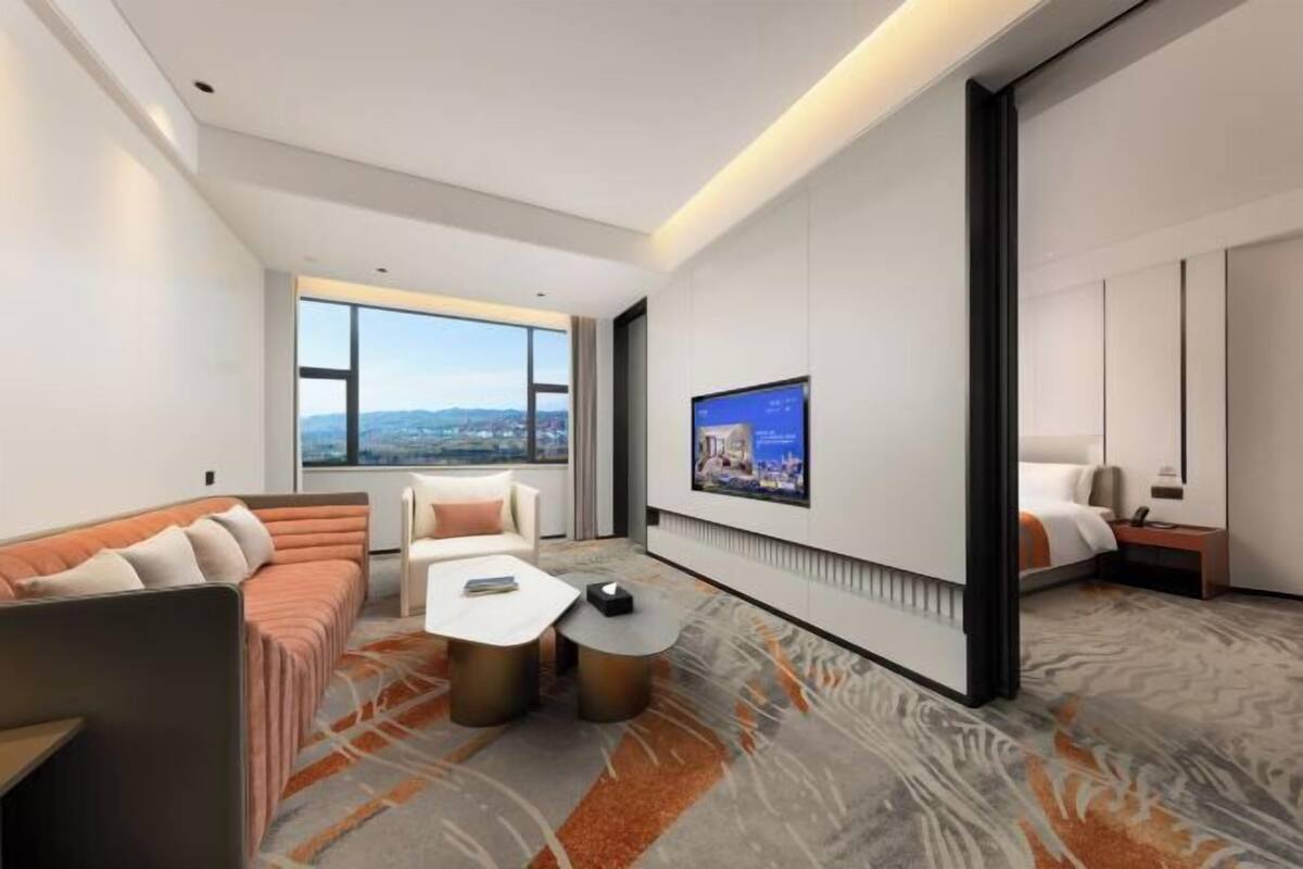 A modern hotel room with a large window, a flat-screen TV, a sofa, a coffee table, and a bed.