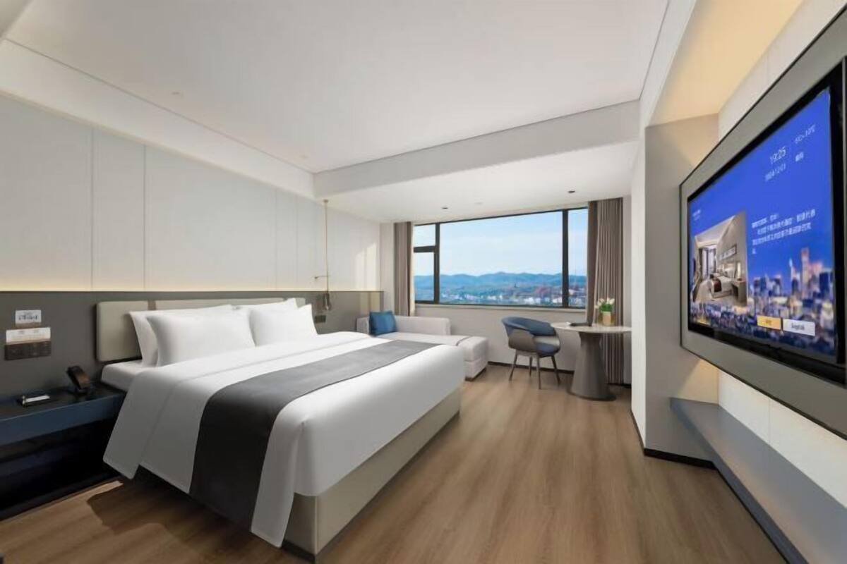 A modern hotel room with a large bed, a desk, a chair, a TV, and a city view.