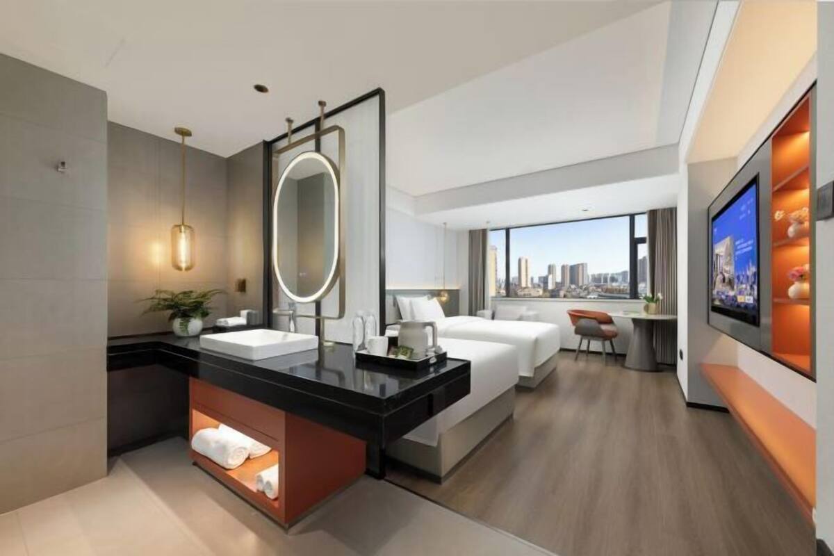 A modern hotel room with a large bed, a separate sitting area, a bathroom with a vanity, and a city view.