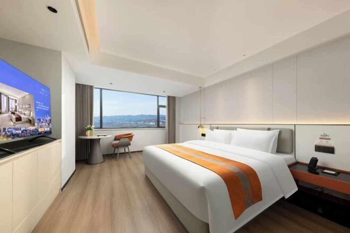 A modern hotel room with a large bed, a desk, a chair, and a TV displaying a cityscape.