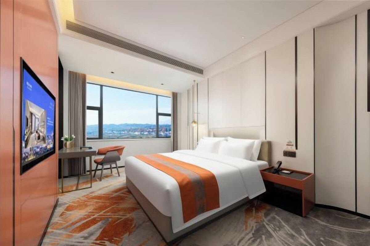 A hotel room with a bed, a desk, a chair, a TV, and a view of the city.