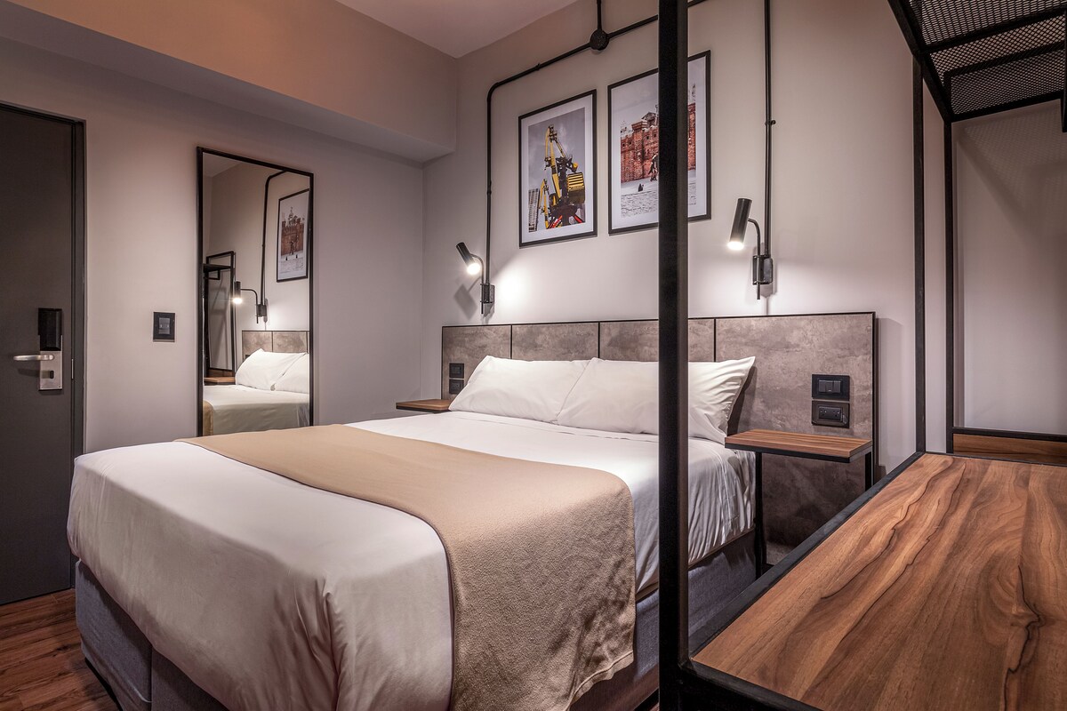 Una habitación de hotel moderna con una cama grande, mesitas de noche, un espejo y cuadros en la pared.