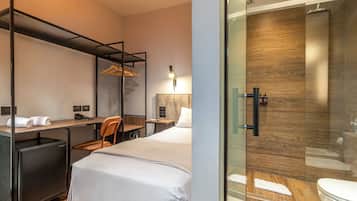 A hotel room with a bed, desk, chair, and a glass-enclosed shower area.