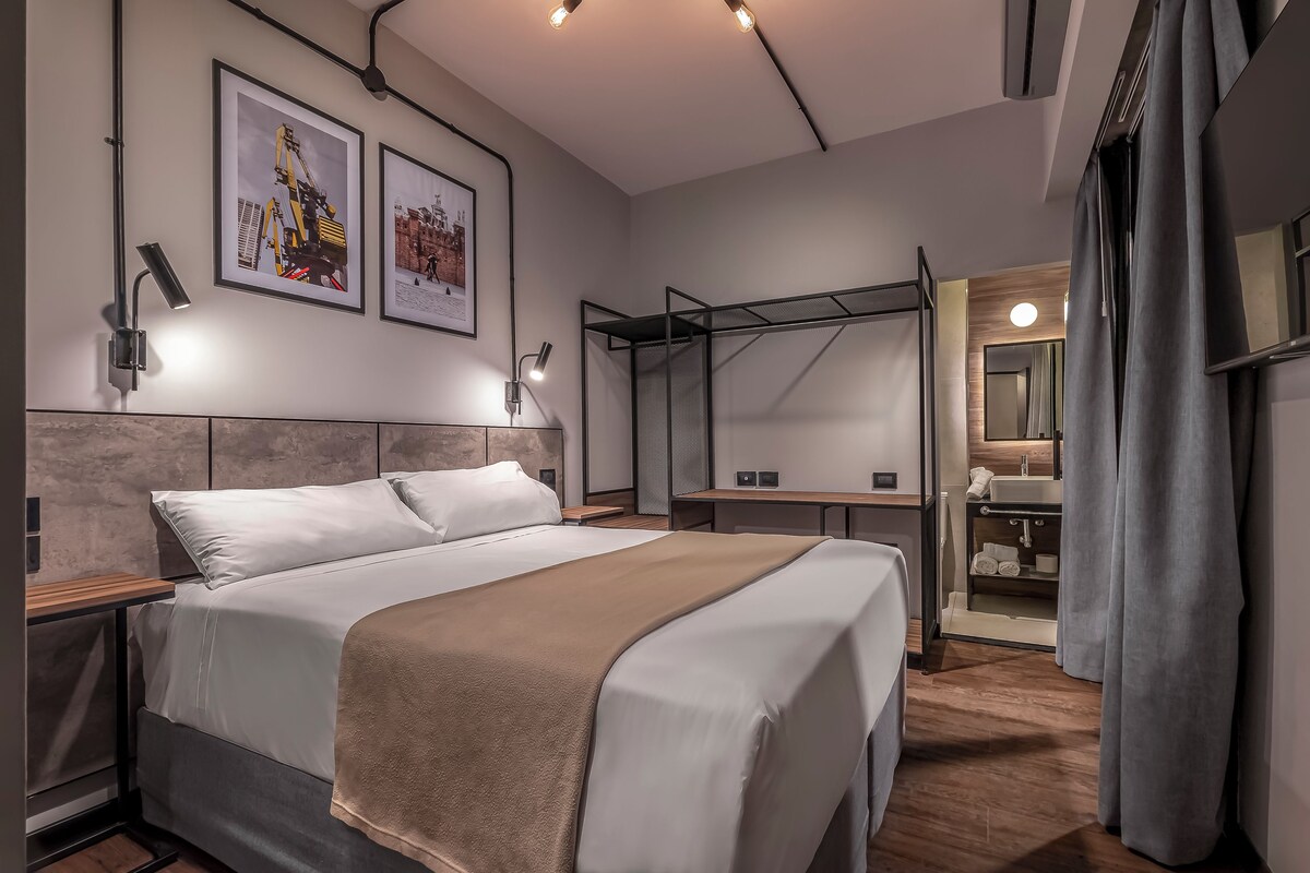 Habitación de hotel moderna con una cama grande, un escritorio y un baño visible a través de un tabique de cristal.