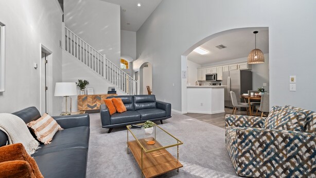 A spacious living area with a dark leather sofa, patterned armchair, and a wooden coffee table. A staircase leads to an upper level, and a kitchen is visible in the background. - 5 Bedroom Spacious Home with Private Pool and Resort Pool in Terra Verde Resort (Kissimmee)