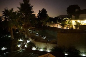 A nighttime view of a swimming pool area with surrounding palm trees and a lit-up building in the background.