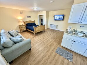 A bedroom with a wooden bed, a grey sofa, a small bedside table, and a lamp. There is a TV mounted on the wall and a kitchen area with a sink and cabinets.