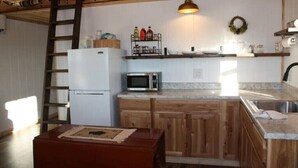A compact kitchen with wooden cabinets, a white refrigerator, a microwave, and a sink. There is a wooden bench with a placemat in the foreground.