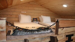 A wooden cabin with a loft bed secured by chains, a small table, and a shelf with a book.