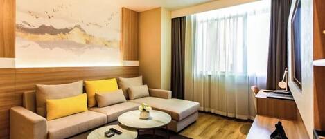 A modern hotel room with a sofa, two coffee tables, a TV, and a large window with curtains.