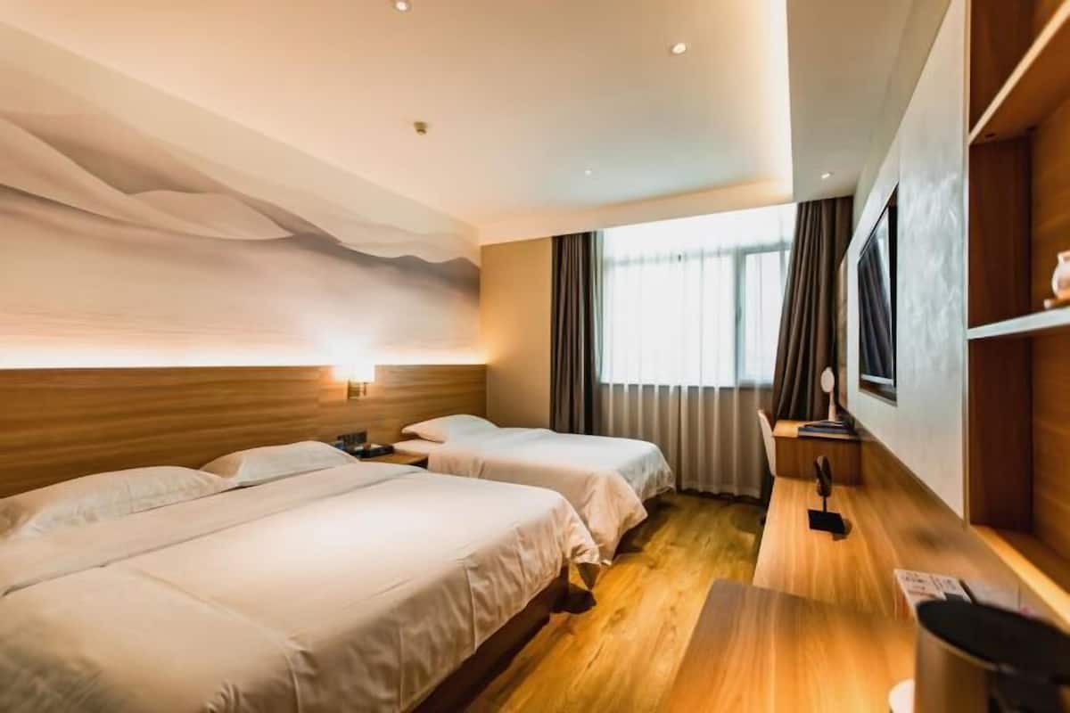 A hotel room with two beds, wooden flooring, a desk, and a large window with curtains.