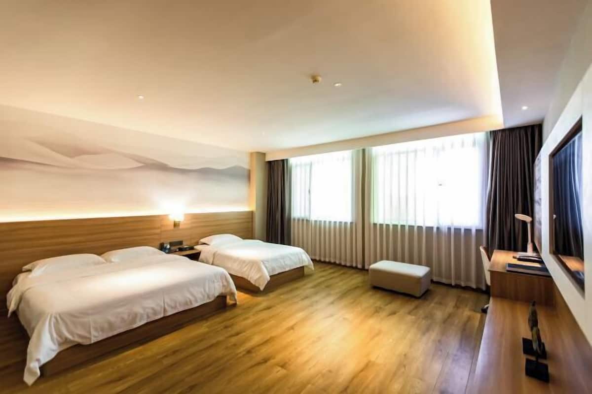 A hotel room with two beds, a wooden floor, a large window with curtains, and a wall-mounted lamp.