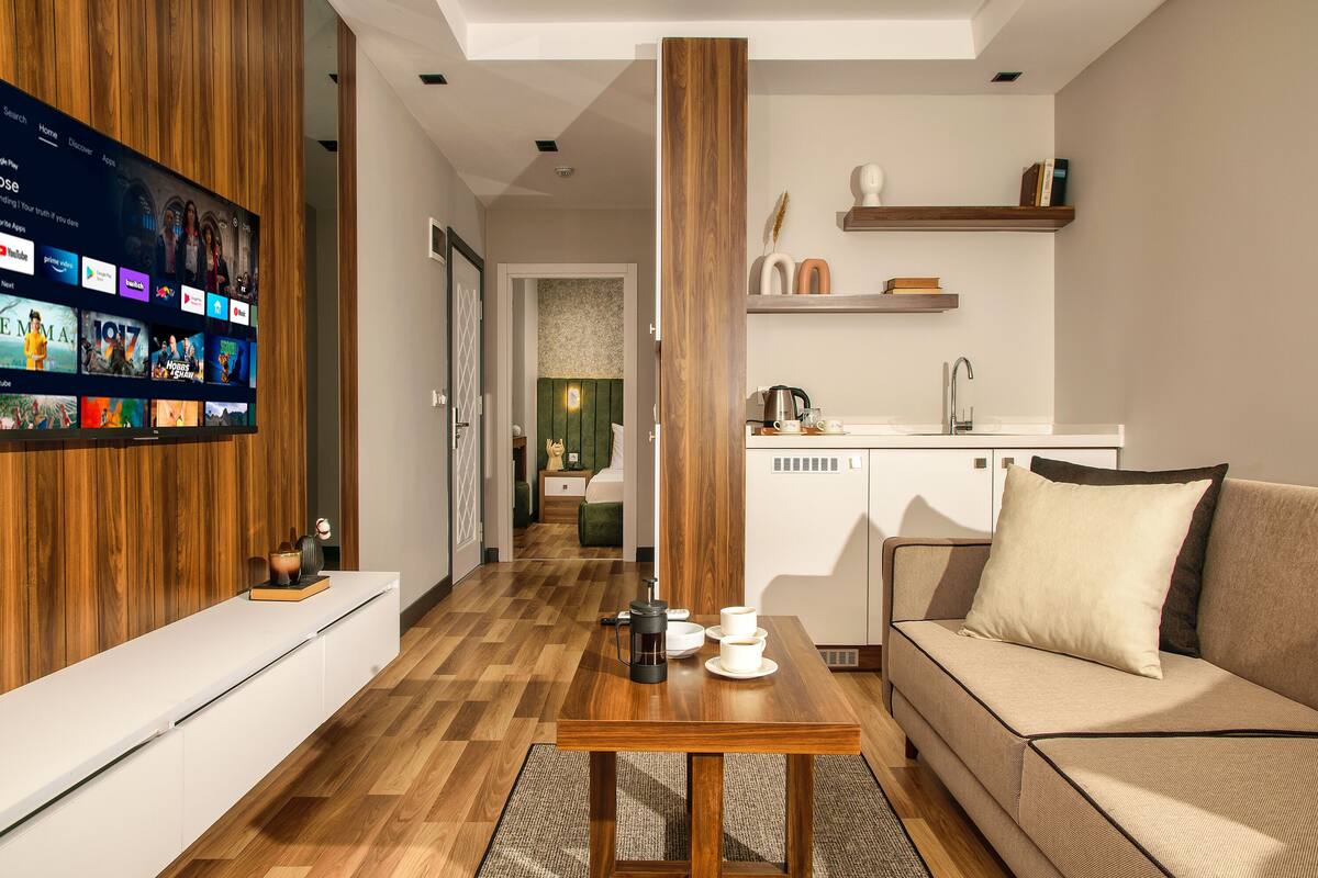 Ein modernes Wohnzimmer mit Flachbildfernseher, Couchtisch aus Holz, einer Couch und einer Küchenzeile mit Spüle und Schränken.