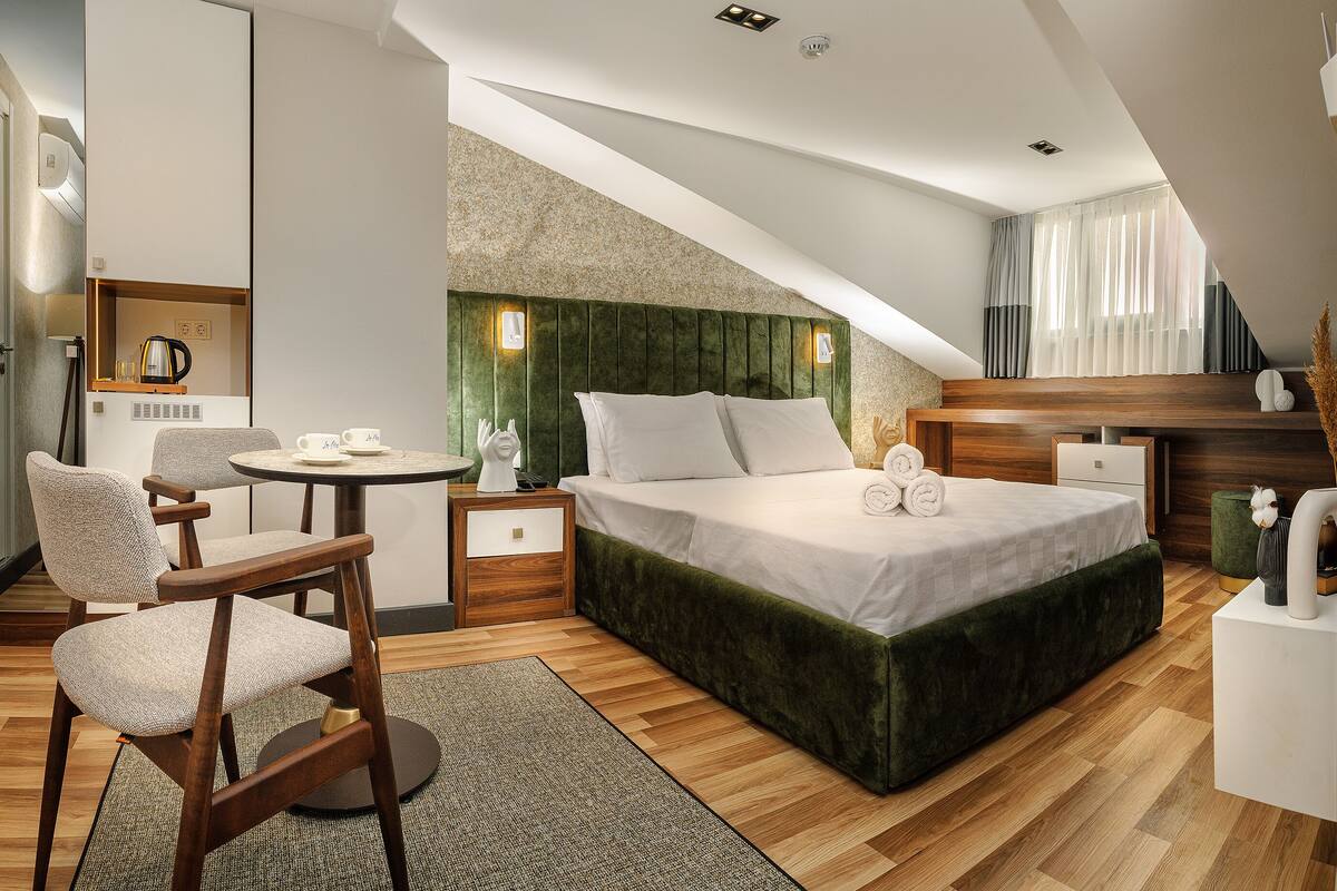 Ein modernes Hotelzimmer mit einem Bett, einem kleinen runden Tisch mit Stühlen, einem Schreibtisch und einem großen Fenster mit Vorhängen.