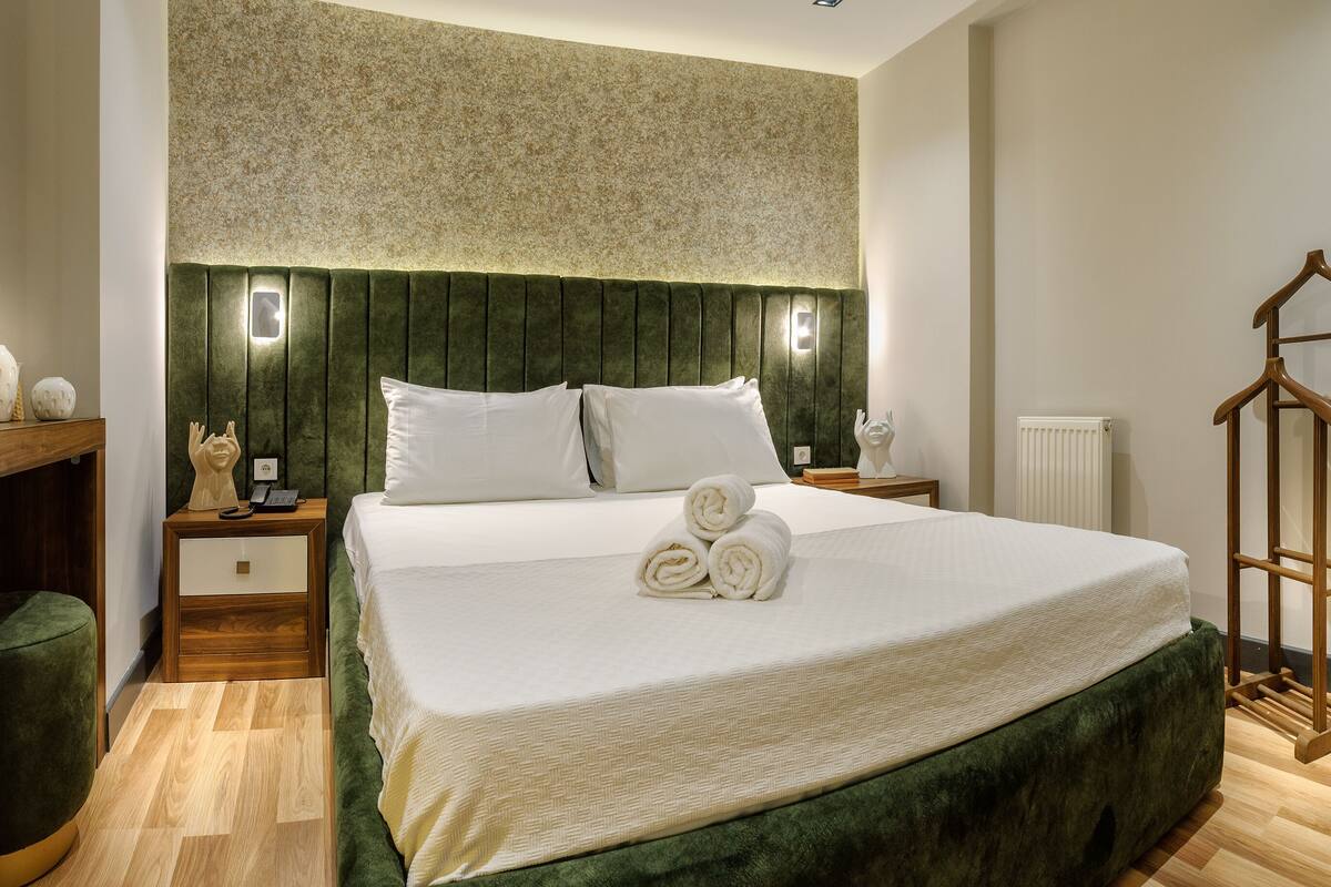 Ein Hotelzimmer mit einem großen Bett, einem hölzernen Nachttisch, einer gepolsterten grünen Bank und einer Holzkommode.