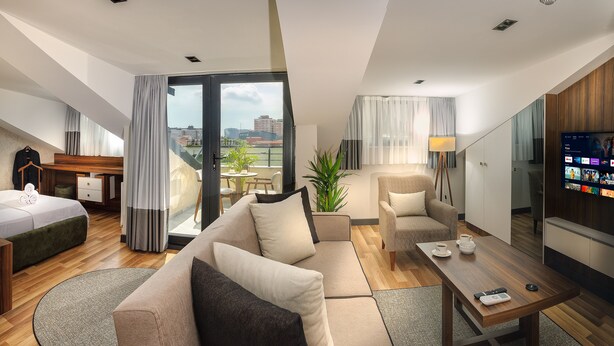 Una habitación de hotel moderna con sofá, sillón y una mesa de comedor. Hay un televisor en la pared y un balcón con vistas a la ciudad.