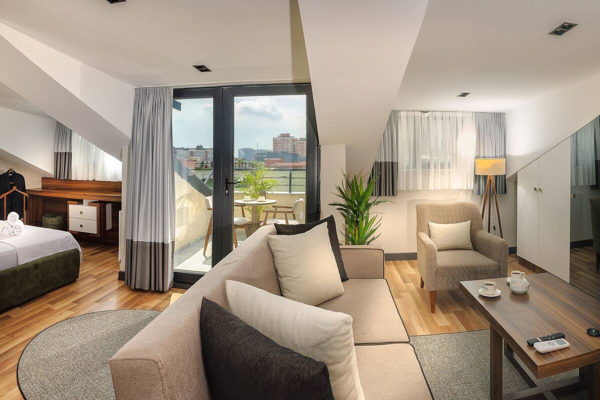 Ein modernes Hotelzimmer mit einer Couch, einem Sessel und einem Esstisch. An der Wand befindet sich ein Fernseher und ein Balkon mit Blick auf die Stadt.