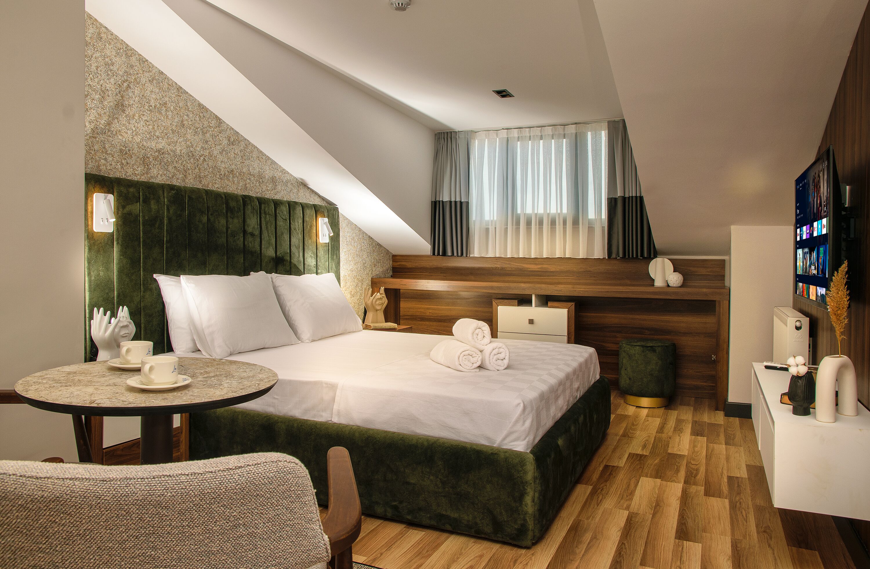 A modern hotel room with a large bed, a round table with a cup, a chair, a small green ottoman, and a TV on a wooden cabinet.
