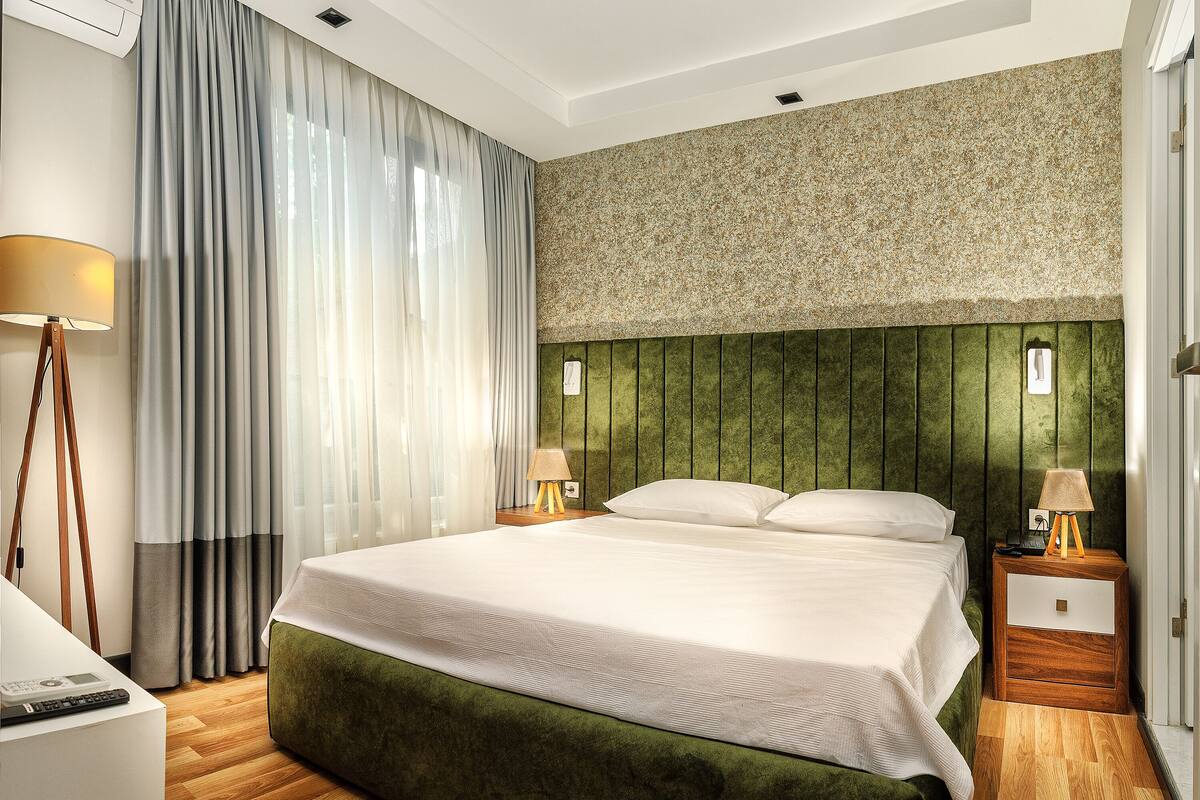 Ein Hotelzimmer mit einem großen Bett, Holzboden, einem grünen, gepolsterten Kopfteil und einer Wand-Klimaanlage.