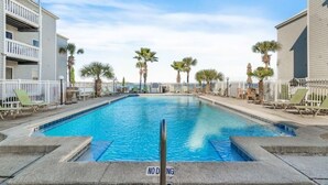 Une piscine rectangulaire, agrémentée d’un panneau indiquant qu’il est interdit de plonger, entourée de chaises longues et de palmiers, avec des bâtiments de chaque côté.