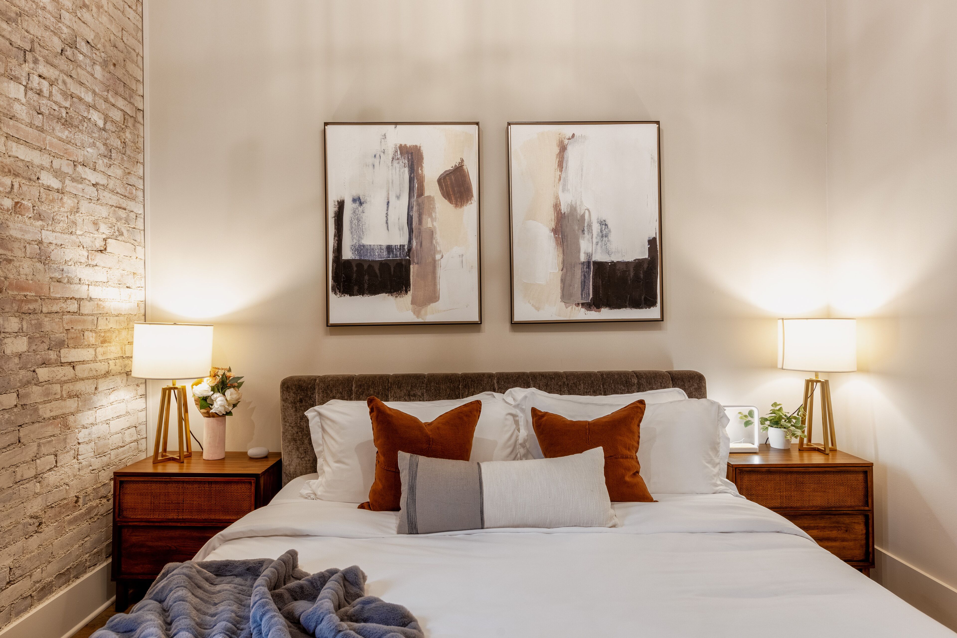 Un lit bien fait, agrémenté d’oreillers blancs et bruns, deux tableaux abstraits accrochés au mur et un mur de briques sur un des côtés.