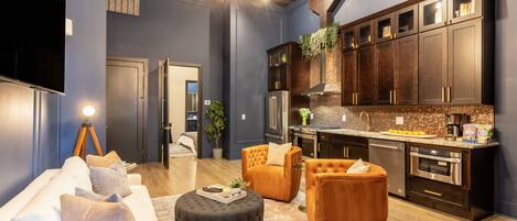 A modern living room with a sofa, ottoman, and chairs, a kitchen with dark cabinets and stainless steel appliances, and a chandelier.
